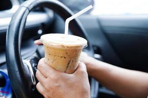 asiatische fahrerin hält eiskaffeetasse zum trinken im auto, gefährlich und riskiert einen unfall. foto