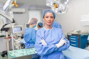 Porträt von lächelnd der Chirurg im Krankenhaus. weiblich Gesundheitswesen Arbeiter ist tragen Peelings. sie ist Stehen mit Waffen gekreuzt gegen Beleuchtung. foto