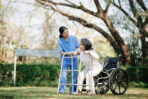 asiatisch Vorsichtig Pflegekraft oder Krankenschwester und das glücklich geduldig im ein Rollstuhl sind Gehen im das Garten zu Hilfe und ermutigen und sich ausruhen Ihre Verstand mit Grün Natur. Hilfe Unterstützung du selber zu lernen zu gehen. foto