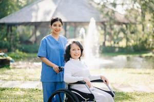 Alten asiatisch Senior Frau auf Rollstuhl mit asiatisch Vorsichtig Betreuer. Pflege- Zuhause Krankenhaus Garten Konzept. im Sonnenlicht foto