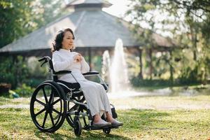 glücklich Alten Frau Sitzung auf Rollstuhl draussen im Park entspannen Ihre Verstand mit Grün Natur. Pflege- Zuhause Krankenhaus Garten Konzept. foto