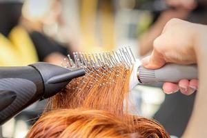 Nahansicht von Meister Hand mit Föhnen und Haarbürste weht weiblich rot Haar im ein Salon. foto