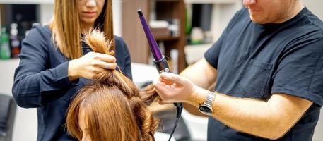 zwei Haar-Stylisten mit ein Eisstockschießen Eisen auf Kunden lange braun Haar im ein Schönheit Salon. foto