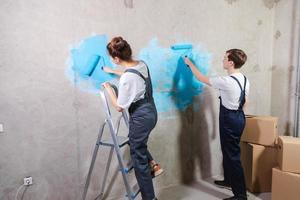 Haus Dekoration DIY Renovierung Konzept. glücklich Paar im leeren Zimmer von Neu Zuhause Gemälde Mauer zusammen. Mann und Ehefrau halten Farbe Walze Gemälde Mauer mit Blau Farbe Farbe im Neu Haus. foto