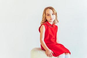 Süss wenig Mädchen im rot Kleid Sitzung auf Stuhl gegen Weiß Mauer beim heim, entspannend im Weiß hell Leben Zimmer drinnen. Kindheit Schulkinder Jugend entspannen Konzept. foto