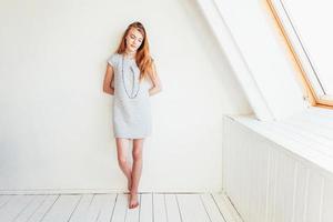 glücklich Teenager Mädchen lächelnd. Porträt jung glücklich positiv Teen Frau im grau Kleid beim Zuhause im hell Zimmer gegen Weiß Mauer. europäisch Frau. positiv Mensch Emotion Gesichts- Ausdruck Körper Sprache. foto