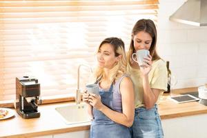 glücklich Lesben Paar halten Tassen von Kaffee im Küche. Paar von Lesben Mädchen genießen Kaffee beim Zuhause nehmen Über etwas. zwei jung Erwachsene schön Frauen trinken Tee im modern Küche. foto