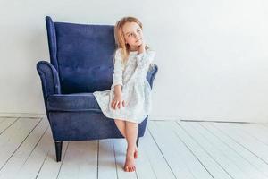 Süss wenig Mädchen im Weiß Kleid Sitzung auf modern gemütlich Blau Stuhl entspannend im Weiß hell Leben Zimmer beim Zuhause drinnen. Kindheit Schulkinder Jugend entspannen Konzept. foto