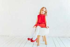 Süss wenig Mädchen im rot Kleid Sitzung auf Stuhl gegen Weiß Mauer beim heim, entspannend im Weiß hell Leben Zimmer drinnen. Kindheit Schulkinder Jugend entspannen Konzept. foto