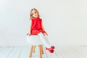 Süss wenig Mädchen im rot Kleid Sitzung auf Stuhl gegen Weiß Mauer beim heim, entspannend im Weiß hell Leben Zimmer drinnen. Kindheit Schulkinder Jugend entspannen Konzept. foto