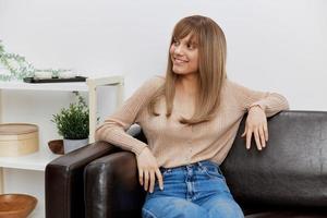 süß lächelnd blond Dame im beiläufig Sweatshirt sieht aus beiseite genießen gut Stimmung Sitzung im Sofa beim modern Leben Zimmer Innere. weiblich Klient haben schrumpfen geplanter Termin im psychologisch Hilfe Büro. Kopieren Raum foto