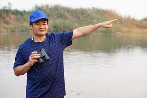 asiatisch Mann trägt Blau Deckel, halt Fernglas und Punkt zu etwas beim See, Natur Quelle. Konzept, Natur Erkundung. Ökologie lernen. Zeitvertreib Aktivität, Lebensstil. Mann erkunden Umgebung foto