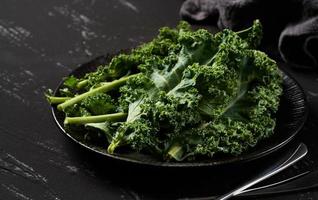 Konzept von frisch Grünkohl Blätter Salat im ein schwarz Gericht auf dunkel Hintergrund. Grün Grünkohl Blätter Salat Essen im das Küche. Löffel, Gabel foto