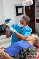 männlich Mitte -alt Zahnarzt beim seine Büro mit ein x Strahl Bild zu erklären Behandlung zu seine Senior Frau geduldig foto