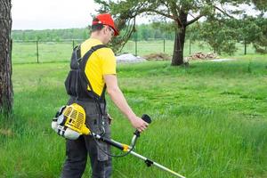 ein männlich Gärtner mäht das Grün Gras von das Rasen im das Hinterhof mit ein Benzin Mäher. Trimmer zum das Pflege von ein Garten Handlung foto