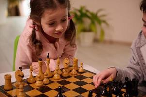 Nahansicht Porträt von ein schön süß wenig europäisch Mädchen spielen Schach Spiel mit ihr ältere Bruder Wer Picks oben ein Schach Stück und machen seine Umzug. intellektuell Clever Entwicklung Tafel Spiele zum Kinder foto