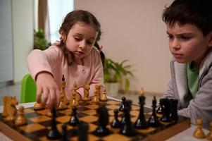 süß wenig Mädchen mit ernst aussehen Sitzung beim das Tabelle und Theaterstücke Schach mit ihr Bruder, pflücken oben ein Schach Stück und Herstellung seine Umzug. früh Entwicklung, Zuhause lehrreich Spiele zum Kinder foto