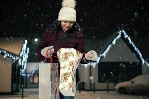 ziemlich Frau mit Einkaufen Taschen Gehen Nieder das Schnee bedeckt Straße auf ein schneebedeckt Winter kalt Abend foto