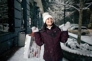 herrlich jung Frau fängt Schneeflocken während Gehen Nieder das Schnee bedeckt Straße mit Einkaufen Taschen foto