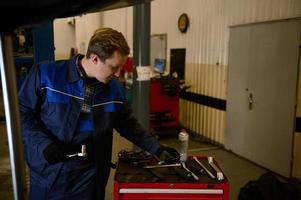 Auto Mechaniker im Uniform mit Arbeit Werkzeug, inspizieren ein angehoben Auto auf ein hissen, während tun Checkliste zum Garantie reparieren Automobil im das Werkstatt Garage foto
