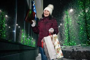 charmant jung Frau Gehen Nieder das Straße mit Einkaufen Taschen mit Weihnachten Geschenk Box im ihr Hände. fröhlich Weihnachten. foto