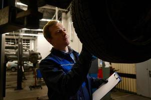 ein jung kompetent Auto Mechaniker inspiziert das Auto angehoben auf das Aufzug zum Schaden. Konzept von technisch Instandhaltung und Auto Bedienung foto