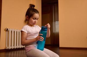 schön kaukasisch wenig Kind Mädchen im Weiß T-Shirt, Sitzung auf Fitness Matte mit ein Blau Sport Flasche von Wasser foto