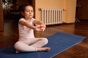 kaukasisch schön Kind Mädchen 5 Jahre alt, Sitzung barfuß auf Yoga Matte, Dehnen Waffen, suchen selbstbewusst beim Kamera foto