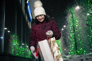 charmant Frau mit Einkaufen Taschen, Gehen Nieder das Straße, aufgehellt durch festlich Erleuchtung beim Winter schneebedeckt Nacht. foto