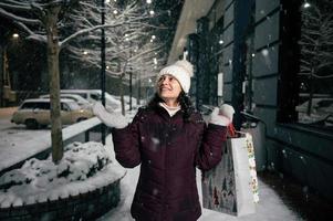 herrlich Frau mit Einkaufen Taschen, fängt Schneeflocken während Spaziergänge entlang ein schneebedeckt Straße beleuchtet durch Urlaub Girlanden foto