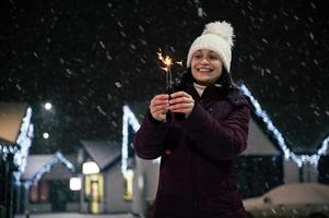 herrlich Frau Beleuchtung Wunderkerzen, Bengalen Beleuchtung auf ein Stadt Straße, beleuchtet durch Girlanden auf ein schneebedeckt Abend foto