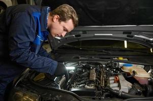 Auto Mechaniker mit ein Taschenlampe Lampe tun ein gründlich Inspektion zum Mängel im das Auto Motor beim Auto Reparatur Geschäft foto