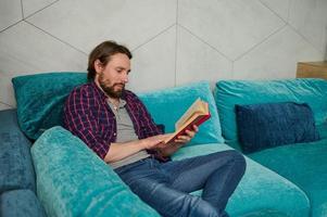 heiter zuversichtlich jung bärtig kaukasisch Mann entspannend auf das Couch, lesen Buch im Zuhause Umfeld. Ausbildung, lernen, Wissen und Gelehrsamkeit Konzept foto