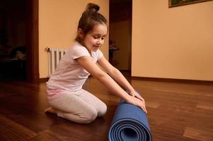 voll Länge schön Vorschulkind Kind Mädchen Sitzung barfuß auf ein hölzern Fußboden und falten Yoga Matte nach Fitness beim Zuhause foto