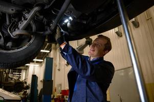 Auto Instandhaltung und Garantie Reparatur im ein Reparatur Geschäft. ein Techniker Verwendet Taschenlampe Lampe Überprüfung das Chassis zum Rad Ausrichtung von ein Auto angehoben auf ein Aufzug im ein Werkstatt Garage. Auto Bedienung foto