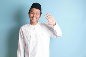 Porträt von asiatisch Muslim Mann im Weiß koko Hemd mit Schädeldecke Hemd Sprichwort Hi, winken Hand beim Kamera, Gruß Person mit glücklich lächeln. isoliert Bild auf Blau Hintergrund foto