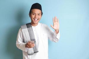 Porträt von jung asiatisch Muslim Mann halten Handy, Mobiltelefon Telefon mit lächelnd Ausdruck auf Gesicht und winken seine Hand, Sprichwort Hallo zu Kamera. isoliert Bild auf Blau Hintergrund foto