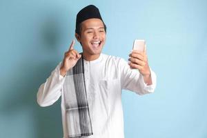 Porträt von asiatisch Muslim Mann im Weiß koko Hemd mit Schädeldecke nehmen Bild von selbst oder Selfie, Sprichwort Hallo und winken seine Hand während Video Forderung. isoliert Bild auf Blau Hintergrund foto
