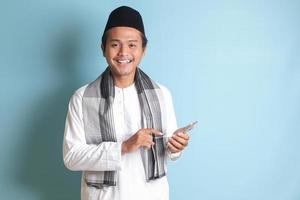 Porträt von jung asiatisch Muslim Mann halten und berühren Handy, Mobiltelefon Telefon mit lächelnd Ausdruck auf Gesicht. isoliert Bild auf Blau Hintergrund foto