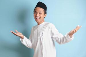 Porträt von asiatisch Muslim Mann im Weiß koko Hemd mit Schädeldecke ziehen Hände gegenüber Kamera, einladend jemand zu Kommen Sie innen, Begrüßung Geste. isoliert Bild auf Blau Hintergrund foto