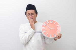Porträt von jung schläfrig asiatisch Muslim Mann Gähnen mit Hand Abdeckung seine Mund während halten ein Uhr Das zeigt an 3 Bin. isoliert Bild auf Weiß Hintergrund foto
