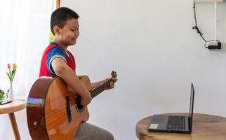 die Geschichte eines Jungen, der einen Notebook-Computer beobachtet, während er sich darauf vorbereitet, zu Hause das Gitarrenspiel zu üben. Jungen nehmen klassischen Gitarrenunterricht online. foto