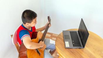 die Geschichte eines Jungen, der einen Notebook-Computer beobachtet, während er sich darauf vorbereitet, zu Hause das Gitarrenspiel zu üben. Jungen nehmen klassischen Gitarrenunterricht online. foto