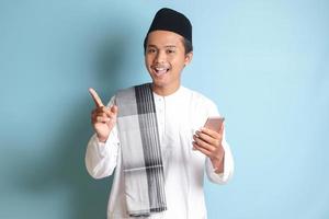 Porträt von jung asiatisch Muslim Mann halten Handy, Mobiltelefon Telefon mit lächelnd Ausdruck auf Gesicht während zeigen Finger zu das Seite. isoliert Bild auf Blau Hintergrund foto