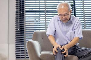 Senior asiatisch Mann Leiden von Knie Arthrose Symptom während Sitzung auf das Couch beim Zuhause mit Kopieren Raum zum medizinisch Chirurgie Behandlung und physisch Therapie Konzept foto
