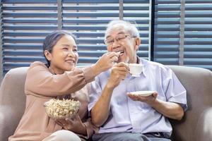 Senior asiatisch Paar ist lächelnd während Sitzung auf das Sofa Couch und haben Spaß Aufpassen Fernsehen mit Popcorn wie Snack zum gut mental Gesundheit und Langlebigkeit Konzept foto