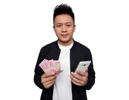 gut aussehend Mann halten Smartphone und halten indonesisch Geld lächelnd suchen beim Kamera foto