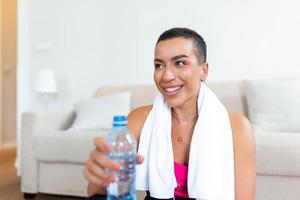 jung Erwachsene afrikanisch amerikanisch Frau Trinken Wasser von Plastik Flasche, Sitzung auf Fitness Matte und ruhen nach Ausbildung beim Zuhause foto