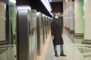 Eine Frau in einer medizinischen Gesichtsmaske wartet auf einen Zug und hält ein Smartphone in der Hand foto