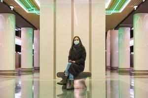 Eine Frau in einer medizinischen Gesichtsmaske wartet auf einen Zug und hält ein Smartphone in der Hand